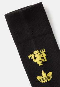 Calzino nero con loghi gialli lavorati a maglia: un leone in cima e un classico trifoglio Adidas in basso, con una superficie liscia.