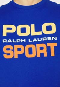 Blå tyg med gul, vit och orange text som läser "POLO RALPH LAUREN SPORT" på en närbild av en crewneck-tröja.