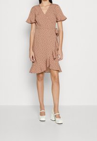 Vestido marrón con lunares, con escote en V, mangas cortas con volantes y un lazo lateral. El dobladillo presenta volantes, combinado con zapatos de tacón bloque blanco.
