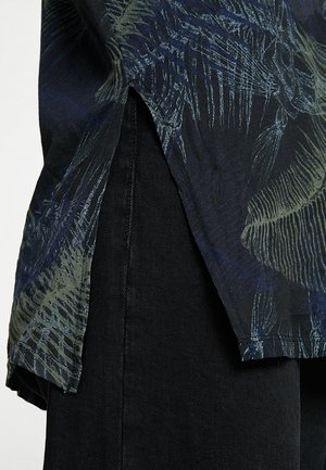 Zwarte shirt met een bladerenpatroon in groene en blauwe tinten, met een gespleten zoom. De stof heeft een gladde textuur. Draagt over donkere broek.