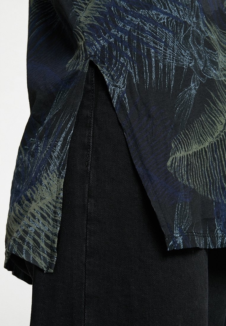 Černá košile s listovým vzorem v zelených a modrých tónech, s rozparky na bocích. Tkanina má hladkou texturu. Nosená přes tmavé kalhoty.