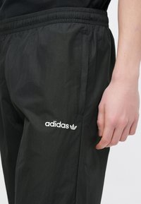 Černé sportovní kalhoty s elastickým pasem a postranními kapsami. Na levé straně mají bílou vyšívanou značku „adidas“ a design s trojlístkem.