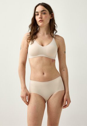 Kvinde med langt brunt hår iført en beige sømløs bh og matchende højtaljede trusser mod en ensfarvet lys baggrund.
