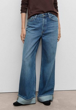 Osoba ubrana w szerokie, podwinięte niebieskie dżinsy, brązową bluzkę z długim rękawem i czarne buty, stojąca na tle gładkiej jasnoszarej ściany.