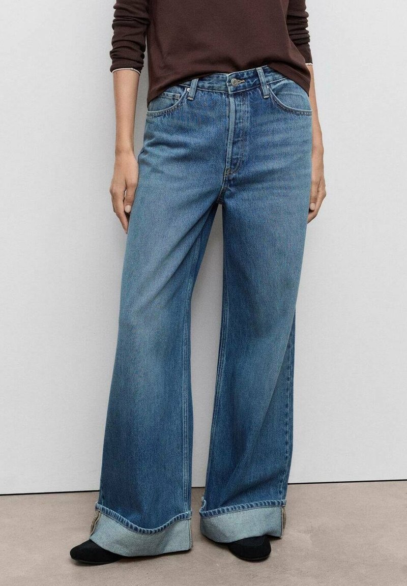 Personne portant un jean bleu à jambes larges et revers, un haut marron à manches longues, et des chaussures noires, debout contre un mur gris clair uni.