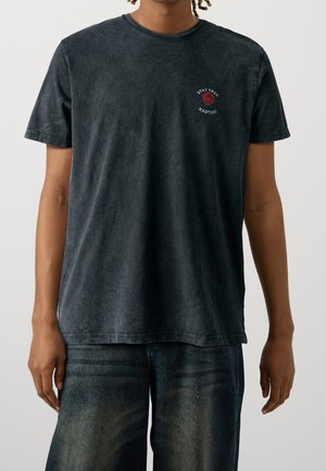 Osoba ubrana w wyblakły czarny T-shirt z małą czerwoną różą i napisem "STAY TRUE KAOTIKO" na górnej lewej klatce piersiowej, w połączeniu z ciemnoniebieskimi dżinsami.