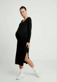 Vestido de maternidade preto com mangas longas e fendas laterais; o detalhamento plissado realça o ajuste. A modelo usa ténis brancos e meias até ao tornozelo.