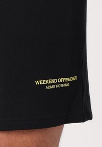 Czarne szorty wykonane z miękkiego materiału, z żółtym napisem "WEEKEND OFFENDER" i "ADMIT NOTHING" po bokach.