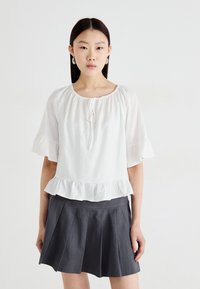 Blouse blanche à manches courtes avec un décolleté froncé et un ourlet festonné. Associée à une jupe plissée grise. Texture lisse, design décontracté.