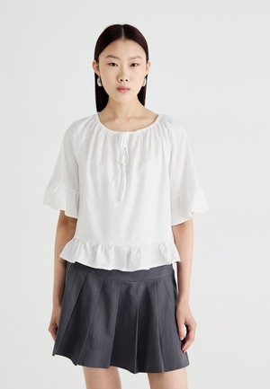 Hvit bluse med korte ermer, samlet halslinning og volangdetalj nederst. Sammen med et grått plisserte skjørt. Jevn tekstur, uformell design.