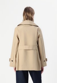 Beige bomullstrenchcoat med bred krage, lös passform och knäppta ärmslut. Har ett bälte baktill och en slät textur.