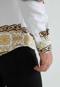 Un bras et une taille d'homme montrant une manche de chemise blanche avec une chaîne en or et des motifs floraux, et un pantalon noir sur un fond uni.
