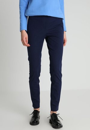 Person trägt dunkelblaue Slim-Fit-Hose, hellblaues Langarmshirt und schwarze glänzende Schnürschuhe und steht vor hellem Hintergrund.