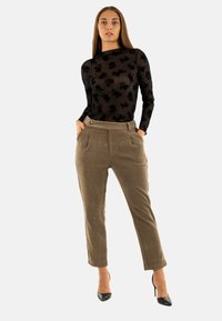 Donna che indossa una blusa nera trasparente a fiori con maniche lunghe, pantaloni in velluto a coste marroni a vita alta e scarpe con tacco nere, posando con le mani nelle tasche.