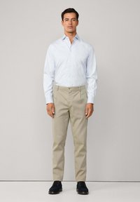 Muškarac stoji u svijetloplavoj kariranoj košulji dugih rukava, bež odijelima i crnim cipelama za odijelo na jednostavnoj pozadini.