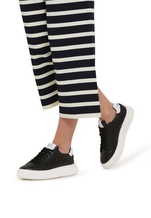 Zwarte sneakers met geometrisch uitsnijdingpatroon, witte rubberen zool en zwarte veters. Gekoeld met zwart-wit gestreepte cropped broeken.