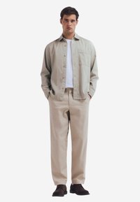 Een lichtgroen overhemd met knopen, een witte T-shirt eronder, beige broek en donkere schoenen. Het overhemd heeft een borstzak.