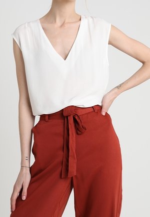 Femme portant un haut blanc sans manches à col en V rentré dans un pantalon taille haute rouge rouille avec une ceinture en tissu nouée en nœud.
