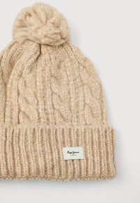 Beige kudumine talve müts pom-pomiga, nööridega kudumise muster, volditud varrukas ja väike ristkülikukujuline kangast silt varruka peal.