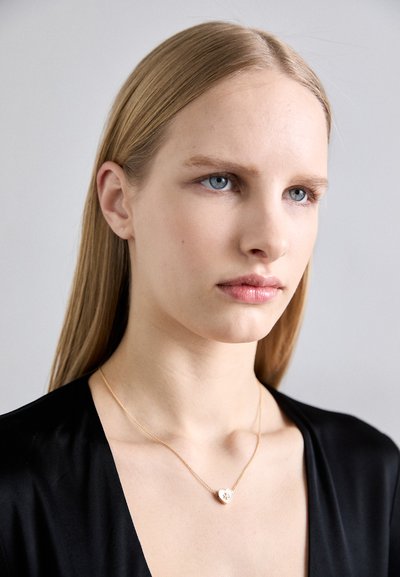 Collier en or avec un pendentif en forme de cœur orné d'un design floral, porté sur le cou d'un modèle contre un fond uni, avec des cheveux lisses.