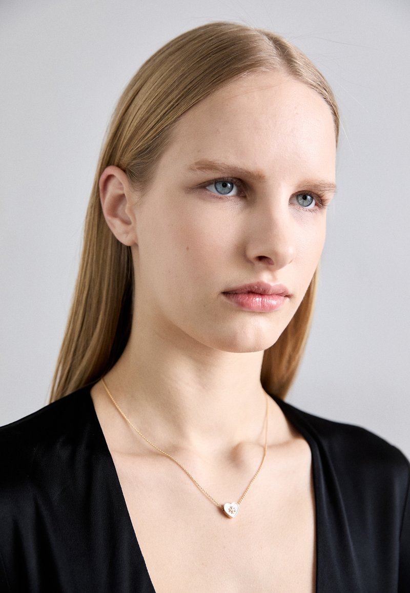 Χρυσό κολιέ με μενταγιόν σε σχήμα καρδιάς που διαθέτει floral σχέδιο, φορεμένο στον λαιμό μοντέλου σε απλό φόντο, με λείο μαλλί.