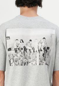 Grå bomulds t-shirt med et stort sort-hvidt grafisk motiv på ryggen, der forestiller seks personer, der sidder på en skyskraber-bjælke over en bylandskab.