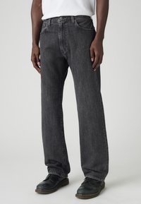 Osoba v volných černých vypraných džínách a černých kožených šněrovacích botách, stojící před světlým jednobarevným pozadím.