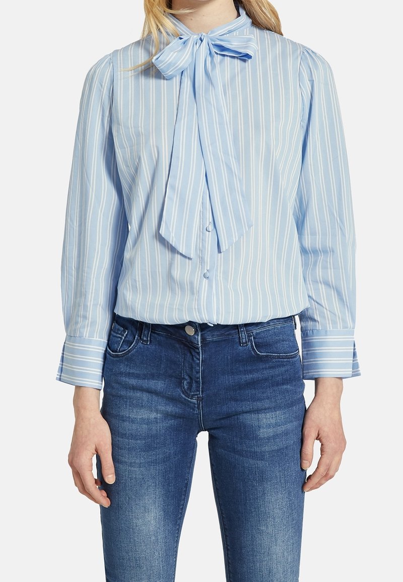 Kvinna i en ljusblå och vit vertikalt randig blus med fluga vid halsen, i kombination med medelblå jeans.