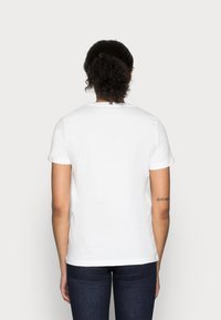 Persona con cabello rizado oscuro recogido en un moño bajo, vistiendo una camiseta blanca de manga corta y jeans oscuros, de espaldas a la cámara.