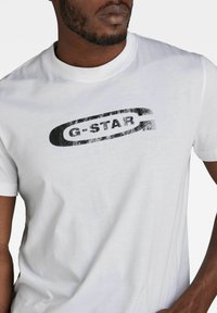 Camiseta blanca de manga corta hecha de algodón, con un logo negro desgastado de "G-STAR" centrado en el pecho. Textura suave, ajuste clásico.