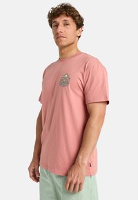 T-shirt en coton rose avec un col rond et des manches courtes, présentant un imprimé graphique coloré sur la poitrine, et une étiquette sur le côté.