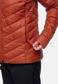 Rostfarbene Puffer-Jacke mit Chevron-Muster, Reißverschluss vorne und verstellbarem Saum. Verfügt über ein grünes Etikett und elastische Kordelzüge.