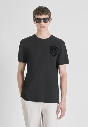 Crni pamučni majica s okruglim izrezom, s teksturiranom crnom logom na lijevoj strani prsnog koša. Model nosi tamne sunčane naočale.