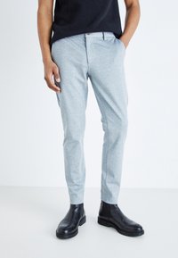 Pantalones slim-fit de color gris claro con una textura sutil, que cuentan con bolsillos laterales y cierre de botón, combinados con botas negras hasta el tobillo.