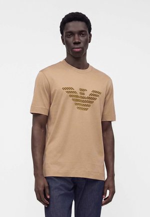 Νέος άνδρας που φοράει ένα μπεζ t-shirt με μεγάλο καφέ λογότυπο αετού και σκούρα μπλε τζιν, στέκεται μπροστά σε απλό λευκό φόντο.