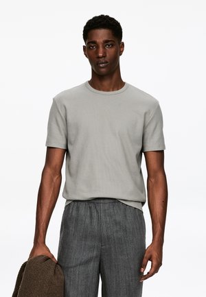 Light gray ribbed short-sleeve t-shirt with a round neckline, paired with gray, textured trousers. Model holds a dark brown knit item.