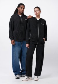 To modeller har på seg overdimensjonerte svarte hettegensere med frontlommer, kombinert med løse jeans og svarte bukser. Omgivelsene er enkle og nøytrale.