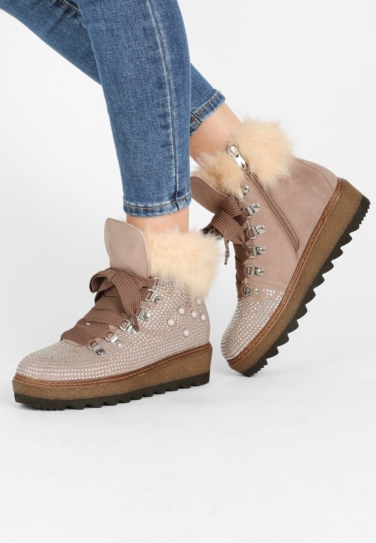 Pieds chaussés de bottines compensées beige avec bord en fourrure, ornements de strass et de perles, lacets marron et œillets métalliques, portées avec un jean bleu.
