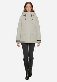Parka impermeabile beige con cappuccio, dotata di tasche frontali, dettagli laterali e polsini elasticizzati. Indossata sopra pantaloni grigi attillati e stivali neri alti.