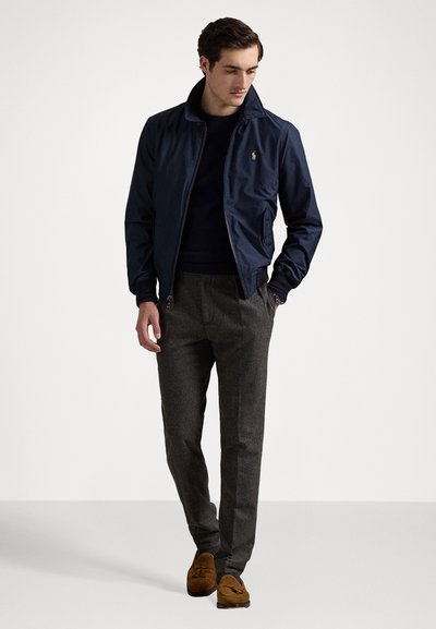 Σκούρο ναυτικό προηγμένο σακάκι με φερμουάρ και όρθιο γιακά, φορεμένο πάνω από ένα σκούρο πουλόβερ. Συνδυάζεται με γκρίζες υφασμάτινες παντελόνες και καφέ μοκασίνια.