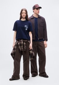 Δύο μοντέλα στέκονται, φορώντας σκούρες φαρδιές παντελόνες και μπλε μπλούζες. Το ένα έχει ένα καρό πουκάμισο δεμένο στη μέση, ενώ το άλλο φορεί καφέ μπουφάν και καπέλο.