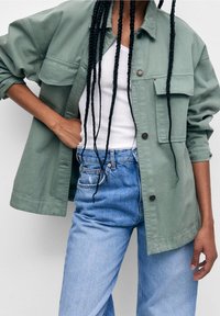 Olive green cotton shirt jacket featuring two front pockets and a relaxed fit, layered over a white tank top and paired with blue jeans.