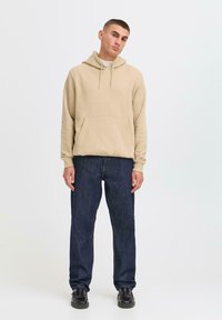 Young man stands confidently, wearing a beige hoodie, dark jeans, and black shoes, against a plain light background.