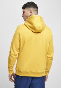 Felpa gialla con tasca a marsupio, cappuccio regolabile con cordini e polsini a coste. Tessuto liscio e design aderente. Abbigliamento sportivo casual.