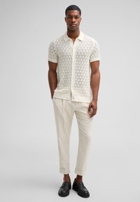 Kurzarmpullover in cremefarbe mit geometrischem Muster, kombiniert mit hellbeigen Hosen und schwarzen Mokassins.