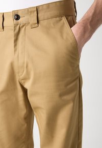 Main droite dans la poche d'un pantalon beige avec un bouton visible et des passants de ceinture, sur un fond clair uni.