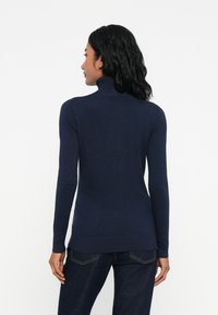 Mujer con cabello oscuro ondulado, vistiendo un suéter de cuello alto azul marino y jeans oscuros, vista desde la espalda sobre un fondo blanco.