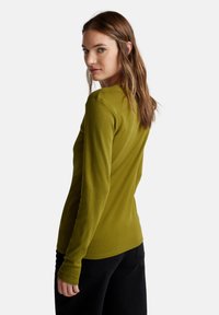 Žena se světle hnědými vlasy, oblečená v olivově zeleném tričku s dlouhým rukávem a černých kalhotách, se dívá přes rameno na bílém pozadí.
