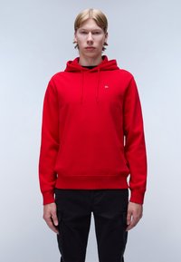 Sudadera roja con capucha ajustable y un pequeño logo en el pecho. El material parece suave con una textura lisa. Combinada con pantalones negros.