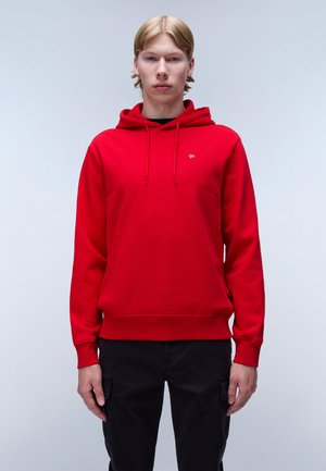 Sweat à capuche rouge avec un cordon de serrage et un petit logo sur la poitrine. Le matériau semble doux avec une texture lisse. Associé à un pantalon noir.